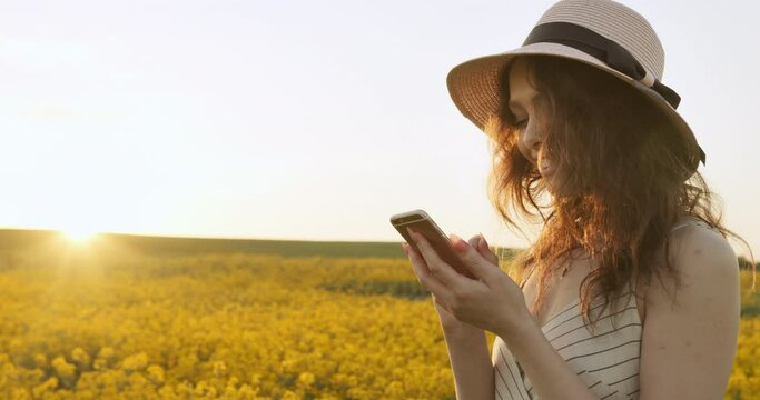 A smiling girl is texting on her phone. Girl in a hat and dress. Sunset in the background. 4k
