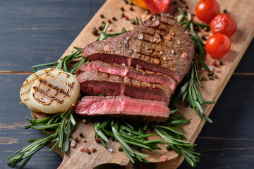 Sliced freshly cooked steak with blood, served with grilled vegetables and fresh rosemary on a wooden board. Close-up.