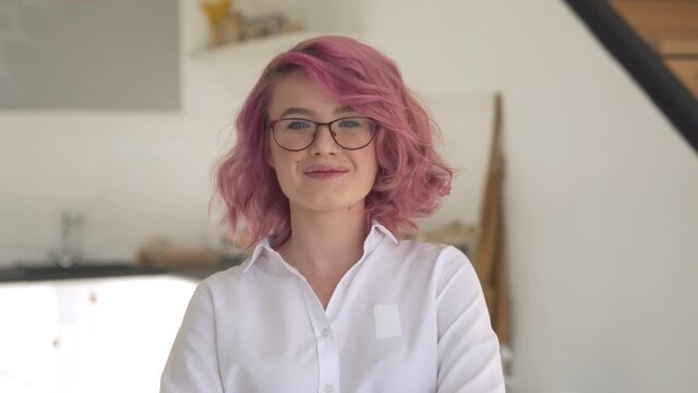 Happy young adult hipster gen z teen girl smiling face with pink hair wearing glasses looking at camera stand in modern kitchen interior. Female small business cafe shop owner entrepreneur portrait.