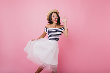 Blissful tanned woman waving hand while dancing in studio. Indoor portrait of pleased brunette girl wears elegant lush skirt.