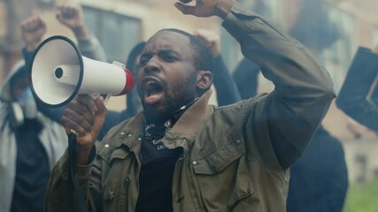 African-American young handsome man screaming in megaphone at protest for human rights outdoors in smoke. Group of people protesting at street. Strike against violence.