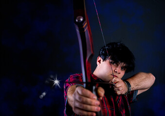 Portrait of a European young man, 21 years old with wolfs tattoo on arm, against a black background, spans a takedown recurved bow and aims
