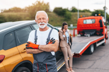 Confident senior man worker in towing service standing in front of his truck with crossed arms and looking at camera.