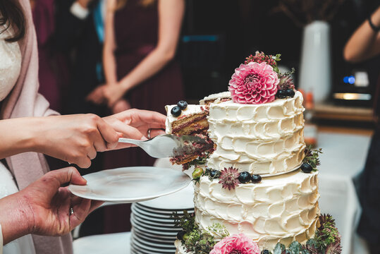 Woman slicing beautiful white wedding cake and serving piece on white plate. Cake decorated with fresh flowers and berries. Wedding day concept.