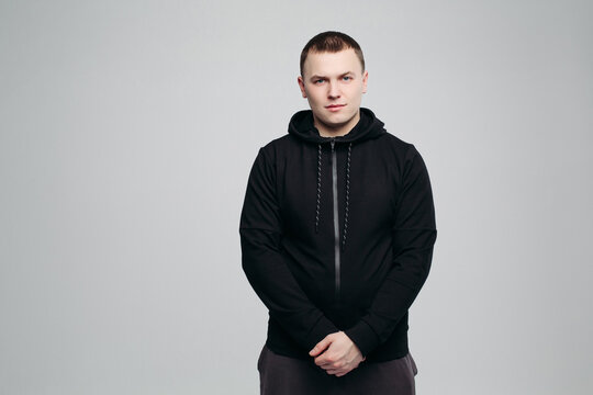 Studio portrait of young, handsome man in black hoodie, standing and posing at camera with hands together. Sporty male in black wear. Man s fashion and male look. Gray background.