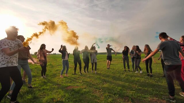 A group of happy friends are running with colored smoke across the field at sunset.