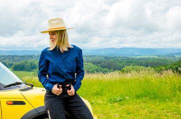 Blonde cowgirl in hat near a car at meadow with mountains on background