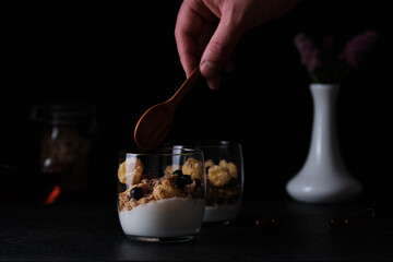 A man takes a spoon with granola and yogurt served in a glass for dinner. Black background around. Hands of a man in a frame.