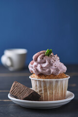 Festive cupcake with pink cream on a plate. The dessert is decorated with mint leaves and chocolate chips. Background color classic blue.