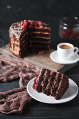 A slice of cake with chocolate and cherry on a plate. A cup of espresso and a cake in the background.