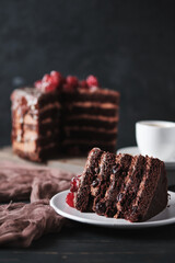 A slice of cake with chocolate and cherry on a plate. A cup of espresso and a cake in the background.
