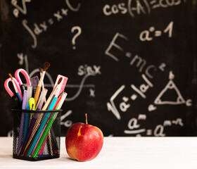 Coloured school supplies and apple on white wooden contertop on chalkboard background with formulas. Education concept