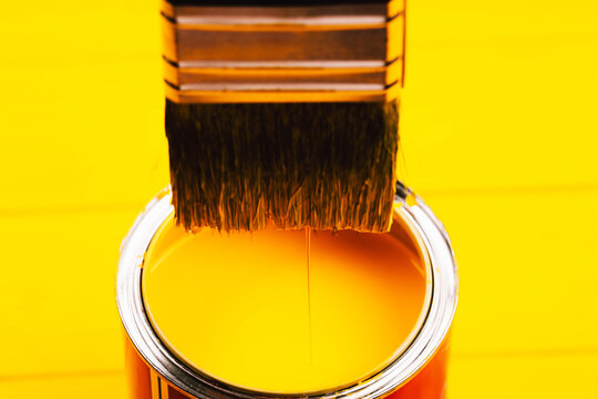 Yellow Paint Is Dripping From Brush Into Open Can On Freshly Painted Wooden Background. Renovation Concept. Macro.