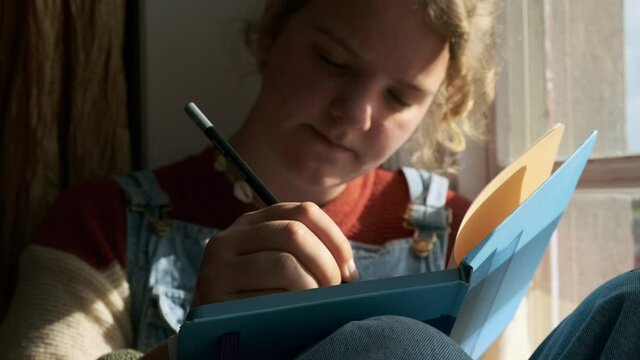  Teenage girl sitting by window and writing in note pad