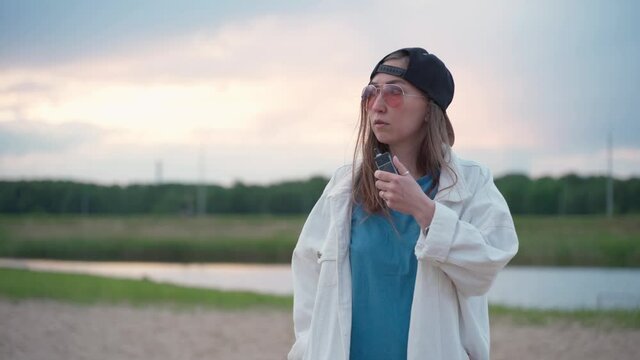Pretty young girl in cap vape popular ecig gadget vaping deviceat the sunset. Happy brunette vaper girl with e-cig. Portrait of smoker female model with electronic cigarette vaporizer. Ejuice vaping