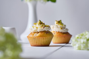 Fruit cupcakes with cream, decorated with ground pistachio and flowers in a vase on a white wooden table. Morning, rustic still life.