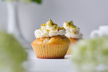 Fruit cupcakes with cream, decorated with ground pistachio and flowers in a vase on a white wooden table. Morning, rustic still life.