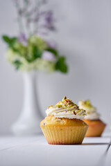 Fruit cupcakes with cream, decorated with ground pistachio and flowers in a vase on a white wooden table. Morning, rustic still life.