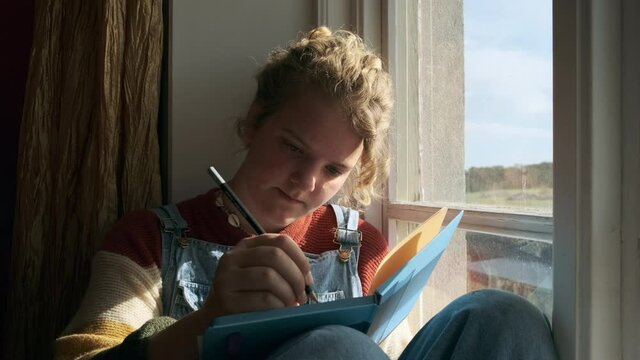  Teenage girl sitting by window and writing in note pad