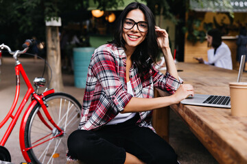 Wonderful girl in good mood sitting on city background with laptop and smiling. Outdoor portrait of attractive brunette lady in glasses posing beside bicycle.