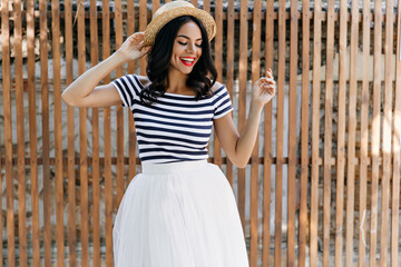 Excited beautiful woman in hat enjoying photoshoot in summer day. Charming tanned female model...