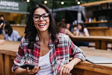 Fashionable caucasian girl with smartphone posing in cafe with smile. Gorgeous female student sitting in outdoor restaurant with laptop.