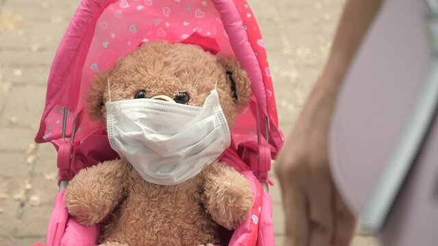 kid on street with his favorite toy in protective mask. healthy childhood concept. little girl walks in park with a pram and a teddy bear in medical mask. child plays an epidemic and protects toy.