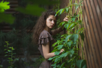 A beautiful, slender girl in a summer sundress poses against a wooden lattice fence with ivy
