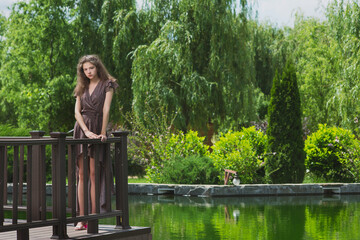 A slender, beautiful girl in a summer sundress on the green shore of an ornamental lake