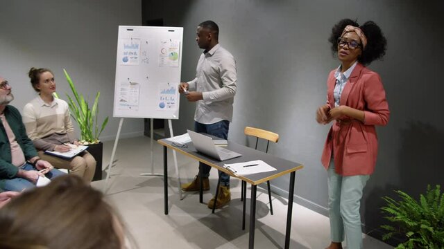 African American male and female business coaches standing in front on mixed-age group of corporate employees, giving a lecture and explaining financial statistics on flipchart