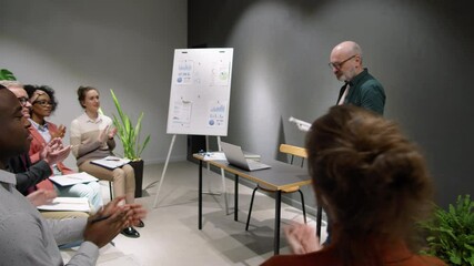Elderly male business coach walking into meeting room while team of diverse people greeting him with applauds, then standing in front of audience, smiling and speaking