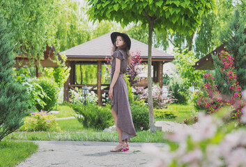 A slender, beautiful girl in a summer sundress and hat poses against the background of nature and wooden houses