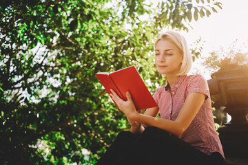 Young beautiful female is thinking about something while is sitting with book in park against green...