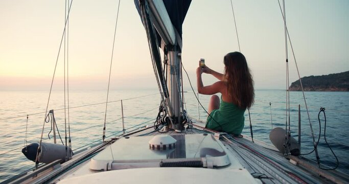 Young woman wearing green sundress enjoying trip on a sailboat in the Ligurian Sea, taking pictures with her mobile phone.