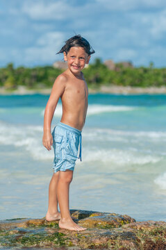 Ni&ntilde;o en traje de ba&ntilde;o azul sonr&iacute;e parado sobre roca en las aguas azules del Mar Caribe.