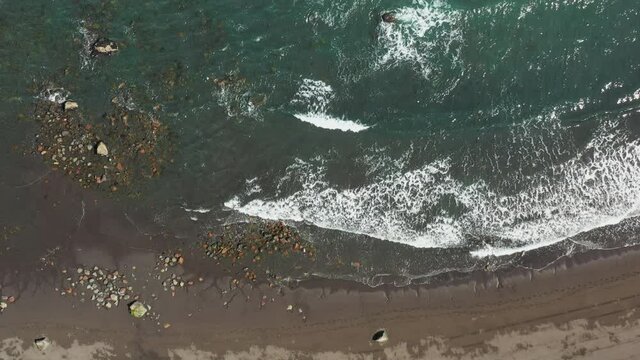 Aerial Overhead View Of Very Shallow Sea Water Over Dark Sandy Sea Floor With Small Waves Washing In. 