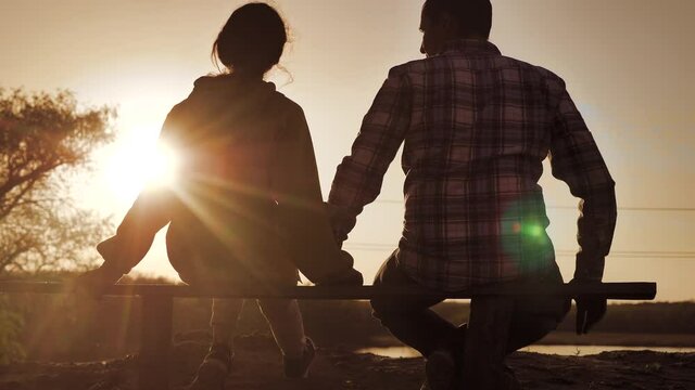 happy family man and girl teamwork sitting on a bench silhouette sunset in lifestyle the park and nature. dad and daughter relax in the park spend together. couple in love hold hand sit back look at