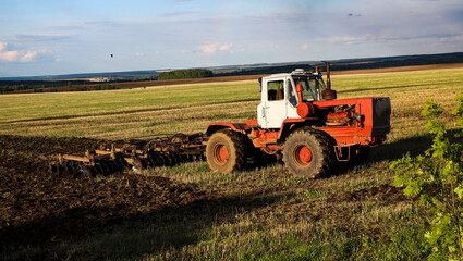 Obraz premium tractor plows and harrows land in large field on sunny spring day. preparing soil for planting crops, plowing soil with tractor with disk plow.