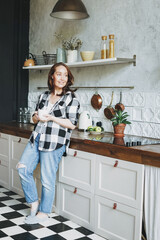 Adult smiling brunette woman in casual plaid shirt and jeans with cup of tea on kitchen at the home