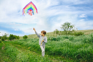 Cute white kid playing colorful kite outdoor at summer meadow or park. Color photography of caucasian young boy isolated at sunny blue sky and green field background