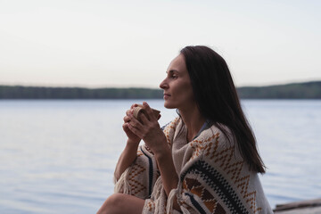 A peaceful beautiful dark haired young woman sits by the lake with a сup of tea and looks into the distance with pleasure
