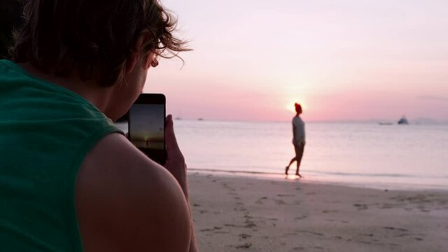 Rear view of a man photographer taking photos of girl walking along the coastline at sunset on the background of the ocean. Model posing for a photographer