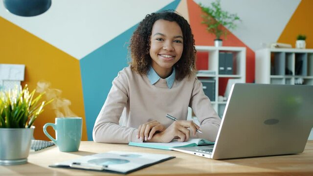 Endless loop portrait of Afro-American girl in office with laptop and coffee cup, woman is looking at camera and smiling while steam is rising from mug.