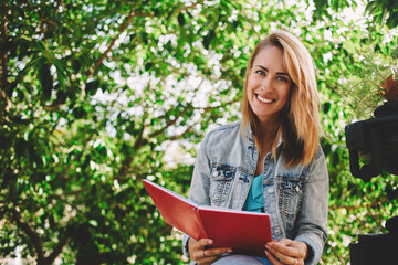 Young beautiful female is thinking about something while is sitting with book in park against green leaves