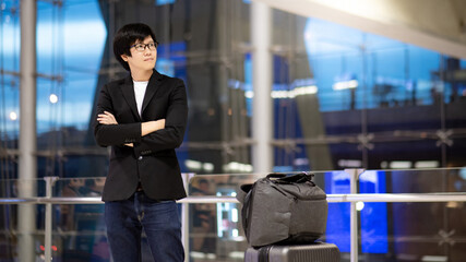 Business travel and baggage claim check-in concept. Smart Asian businessman wearing formal suit jacket standing with suitcase luggage and backpack waiting for flight in international airport terminal.