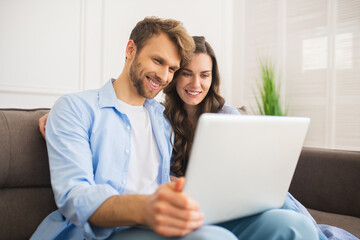 Young couple in denim sitting on a sofa watching something online and smiling