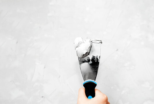 metal spatula with putty or paint with a black handle and a blue stripe in a light skinned hand against a background of light grey concrete surface