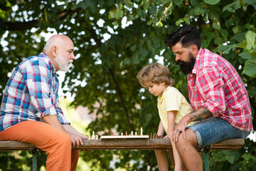 Boy with father and grandfather. Little boy playing chess with his Grandfather and Father. Summer and active holidays. Senior man thinking about his next move in a game of chess.