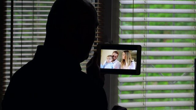 A bald man is facing the light at a window with blinds, she is talking on a video conference on a tablet with a group of different people: a woman mom, a man dad and a little girl.