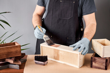 Man carpenter varnishing wooden crate for flowers with brush in her small business woodwork workshop. In your work, do you use stains or wood preservatives to show the wood pattern.